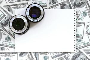 dos lentes fotográficos y un cuaderno blanco se encuentran en el fondo de muchos billetes de dólar. espacio para texto foto