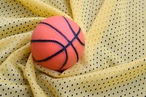 Small orange rubber basketball lies on a yellow sport jersey clothing fabric texture and background with many folds photo