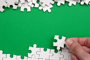 The hand folds a white jigsaw puzzle and a pile of uncombed puzzle pieces lies against the background of the green surface. Texture photo with space for text
