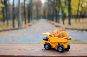 A small toy yellow truck is loaded with yellow fallen leaves. The car stands on a wooden surface against a background of a blurry autumn park. Cleaning and removal of fallen leaves. Seasonal works photo