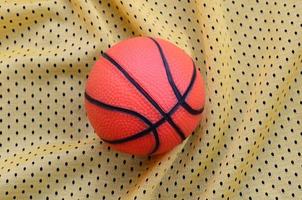 Small orange rubber basketball lies on a yellow sport jersey clothing fabric texture and background with many folds photo