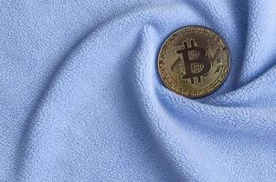 The golden bitcoin lies on a blanket made of soft and fluffy light blue fleece fabric with a large number of relief folds. The shape of the folds resembles a fan from a video card cooler photo