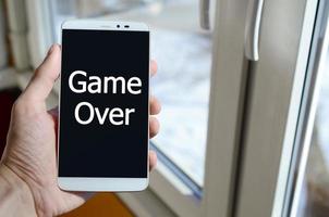 A person sees a white inscription on a black smartphone display that holds in his hand. Game over photo