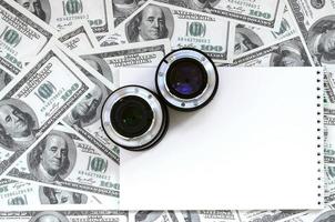 Two photographic lenses and white notebook lie on the background of a lot of dollar bills. Space for text photo