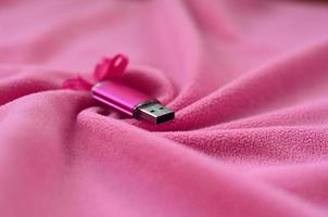 la tarjeta de memoria flash usb de color rosa brillante con un lazo rosa yace sobre una manta de tela suave y peluda de color rosa claro con muchos pliegues en relieve. dispositivo de almacenamiento de memoria en diseño de mujer foto