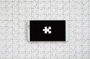 A modern big smartphone with several puzzle elements on the touch screen lies on a white jigsaw puzzle in an assembled state photo