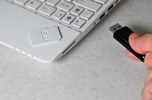 A male hand inserts a black compact USB card into the corresponding input in the side of the white netbook. Man uses modern technologies to store memory and digital data photo