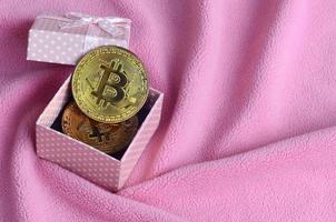 The golden bitcoin lies in a small pink gift box with a small bow on a blanket made of soft and fluffy light pink fleece fabric with a large number of relief folds photo