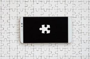 A modern big smartphone with several puzzle elements on the touch screen lies on a white jigsaw puzzle in an assembled state photo