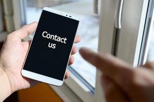 A person sees a white inscription on a black smartphone display that holds in his hand. Contact us photo