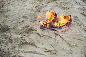 Burning sports sneakers or gym shoes on fire stand on sandy beach coast. Athlete burned out. Physical exertion during training concept photo