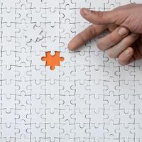 The texture of a white jigsaw puzzle in the assembled state with one missing element, forming an orange space, pointed to by the finger of the male hand photo