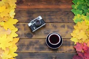 una taza de té y una cámara vieja entre un conjunto de hojas de otoño caídas amarillentas sobre una superficie de fondo de tablas de madera natural de color marrón oscuro foto