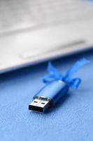 Brilliant blue usb flash memory card with a blue bow lies on a blanket of soft and furry light blue fleece fabric beside to a white laptop. Classic female gift design for a memory card photo