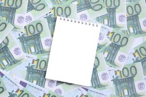 White notebook with clean pages lying on a set of green monetary denominations of 100 euros. A lot of money forms an infinite heap photo