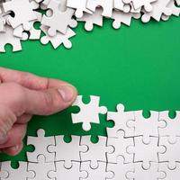 The hand folds a white jigsaw puzzle and a pile of uncombed puzzle pieces lies against the background of the green surface. Texture photo with space for text