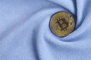 The golden bitcoin lies on a blanket made of soft and fluffy light blue fleece fabric with a large number of relief folds. The shape of the folds resembles a fan from a video card cooler photo