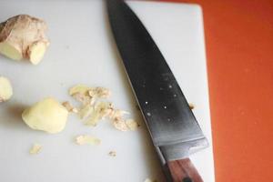 Ginger is being sliced on a plastic chopping board with a knife on an orange table. photo