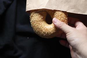 A man's hand is holding a bagel out of a brown paper bag. photo