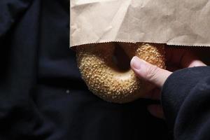 A man's hand is holding a bagel out of a brown paper bag. photo