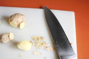 Ginger is being sliced on a plastic chopping board with a knife on an orange table. photo