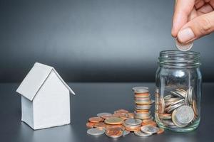 Hand putting Thailand coins in jar with heap coins on black background with house model photo