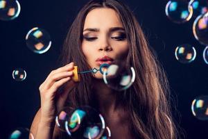 Bubble fun. Beautiful young woman blowing bubbles while standing against black background photo