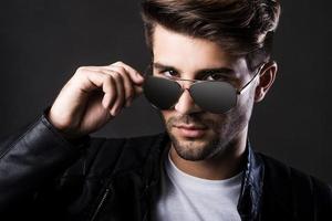 Trendy look. Handsome young man adjusting his sunglasses and looking at camera while standing against black background photo