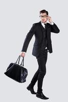 Perfect style. Full length of confident young man in formalwear and eyeglasses carrying briefcase while walking against grey background photo