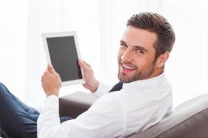 Happy with his new gadget. Rear view of confident young businessman in shirt and tie holding digital tablet and smiling while sitting at the chair photo