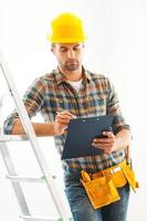 maestro de la construcción en el trabajo. un joven constructor serio escribiendo en el portapapeles y apoyándose en la escalera mientras está de pie en el interior foto