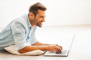 Surfing the net at home. Side view of young man working on laptop while lying on the floor at his apartment photo