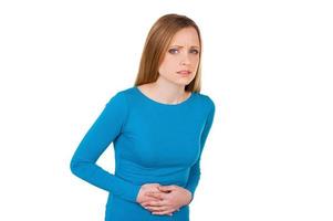 Feeling stomachache. Frustrated young woman holding hands on stomach and looking at camera while standing isolated on white photo