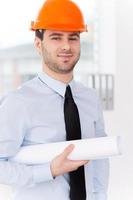 Confident architect. Confident young man in formalwear and hardhat holding blueprint while standing against a window photo