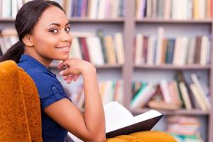 leyendo un libro en la biblioteca. vista trasera de una hermosa estudiante africana sosteniendo un libro y mirando por encima del hombro mientras se sienta en la silla de la biblioteca foto