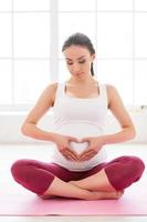 Love and care. Beautiful pregnant woman sitting in lotus position on exercise mat and touching abdomen photo