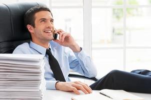 Good business talk. Happy young man in shirt and tie talking on the mobile phone and smiling while sitting at his working place photo