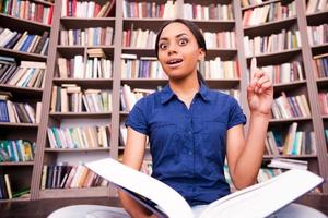 solo inspirado estudiante africana sorprendida sosteniendo un libro y señalando hacia arriba mientras estaba sentada en el suelo en la biblioteca foto