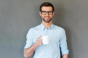 Good coffee for starting your day. Handsome young man in shirt in eyewear holding cup of coffee and smiling at camera while standing against grey background photo