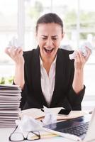 Sick and tired. Furious young woman in formalwear holding papers in hands and shouting while sitting at her working place photo