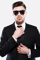 Young and successful. Portrait of handsome young man in formalwear and sunglasses adjusting his necktie and looking at camera while standing against grey background photo