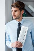 Lost in business thoughts. Thoughtful young man in shirt and tie holding newspaper and looking through a window while standing in office photo