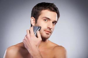 Man shaving. Handsome young man shaving his face with electric shaver and looking at camera while standing isolated on grey background photo