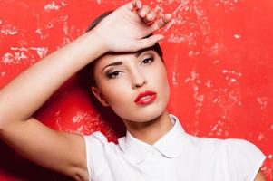 belleza con peinado elegante. vista lateral de una hermosa joven de cabello corto con camisa blanca sosteniendo la mano en la barbilla mientras se enfrenta a un fondo rojo foto