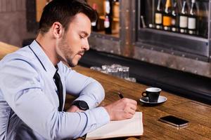 Writing down his fresh ideas. Side view of confident young man in shirt and tie writing something in note pad while sitting at the bar counter photo