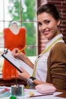 Young and creative. Side view of beautiful young woman drawing fashion sketch and smiling at camera while sitting at her working place with mannequin standing in the background photo