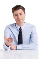 Cheerful interviewer. Handsome young man in shirt and tie sitting at the table and gesturing while isolated on white photo