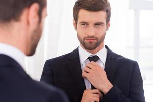 Looking just perfect. Handsome young man in formalwear adjusting his necktie and smiling while standing against mirror photo