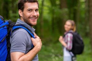 I love traveling. Handsome young man with backpack looking over shoulder and smiling while walking through a forest with woman in the background photo