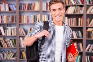 Smart and confident student. Handsome young man holding books and smiling while standing in library photo